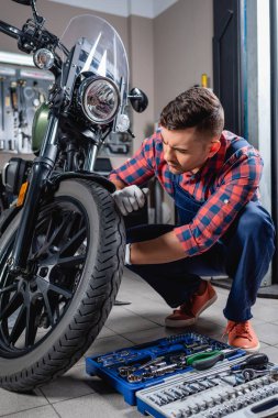 Tulumlu genç tamirci atölyedeki alet çantasının yanında motosiklet kontrolü yapıyor.
