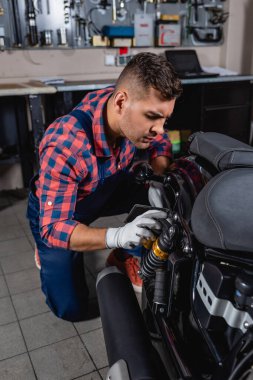 young mechanic in plaid shirt checking shock absorber of motorbike in workshop clipart