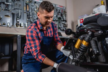 young mechanic in overalls and plaid shirt checking shock absorber of motorcycle in workshop clipart
