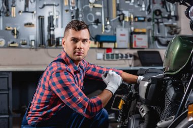 young mechanic looking at camera while examining motorcycle with flashlight clipart