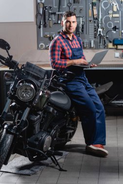 Motosikletin yanında dizüstü bilgisayarla kameraya bakarken tulumlu bir tamirci.
