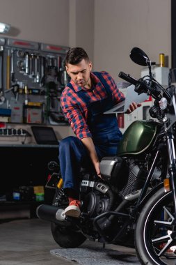 Dizüstü bilgisayar tutarken motorsikleti kontrol eden tulumlu bir tamirci. 