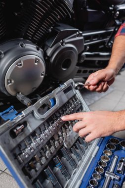 cropped view of mechanic pointing with finger at bits for socket wrench in toolbox near motorbike gearbox clipart