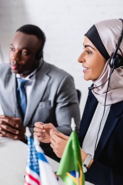selective focus of happy arabian businesswoman in headset near african american business partner on blurred background clipart