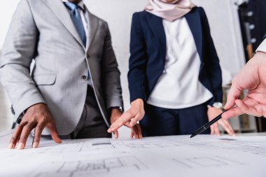 Kırpılmış iş adamı görüşü, bulanık arka planda ırklar arası iş ortaklarının yakınındaki planlara kalemle işaret ediyor.