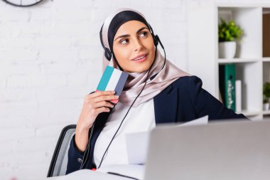 smiling arabian interpreter in headphones holding digital translator with uae flag symbol, blurred foreground clipart