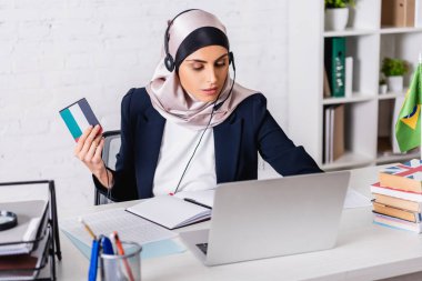 arabian interpreter in headset holding digital translator while working on laptop near dictionaries, blurred foreground clipart