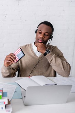 Kulaklık takmış düşünceli Afrikalı Amerikalı tercüman elinde ABD bayrağı amblemi, defter ve dizüstü bilgisayarla ön planda bulanık bir şekilde dijital tercüman tutuyor.