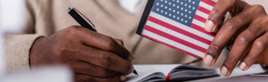 selective focus of african american interpreter writing in notebook while holding digital translator with usa flag emblem, cropped view, banner clipart