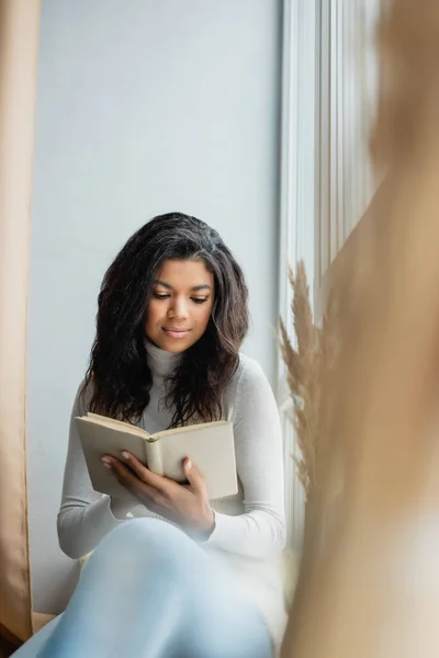 Young African American Woman Reading Book Window Home Blurred Foreground — Stock Photo, Image