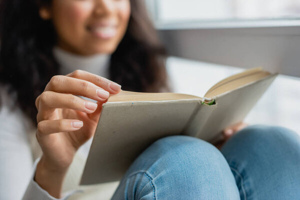 cropped view of african american woman reading book at home, blurred background