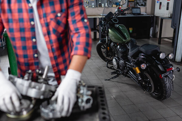 selective focus of motorcycle near mechanic and disassembled gearbox on blurred foreground