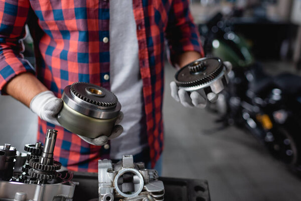 partial view of mechanic in plaid shirt and gloves holding gearwheels of motorbike transmission, blurred background