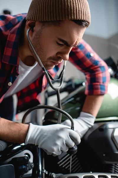 Bereli genç tamirci ve eldivenler steteskoplu motosiklet motorunu inceliyor, geçmişi bulanık.