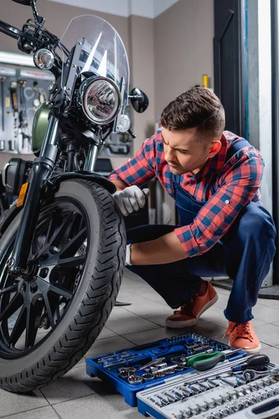 Tulumlu genç tamirci atölyedeki alet çantasının yanında motosiklet kontrolü yapıyor.