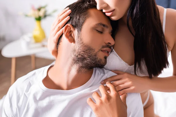 Gözleri kapalı genç bir adam iç çamaşırlı bir kadına yaslanıyor. Yatak odasındaki bulanık arka planda duruyor.