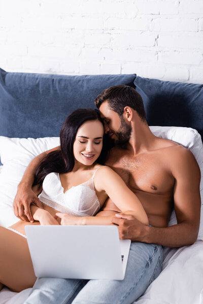 smiling shirtless man hugging woman in lingerie on bed near laptop