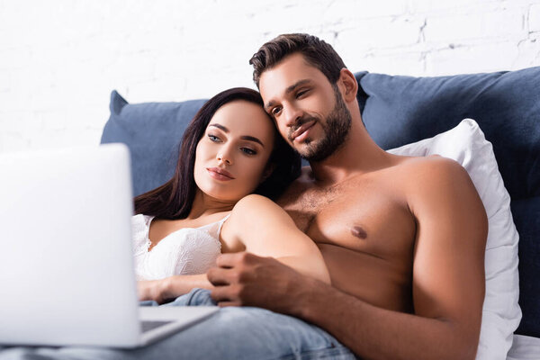 young sexy couple looking at blurred laptop on foreground in bedroom