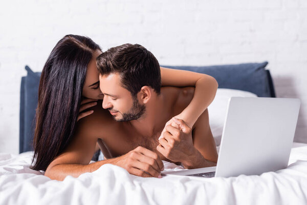 young brunette woman hugging shirtless man near laptop on blurred background in bedroom