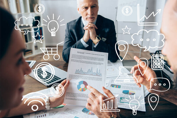 Бизнесмены с бумагами на размытом переднем плане, указывающими на инвестора рядом с ноутбуком в офисе, графиками и иллюстрациями на долларах 