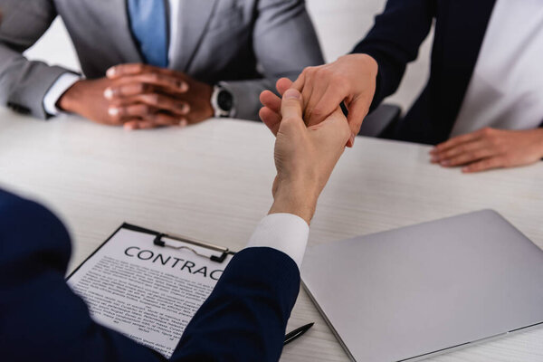 selective focus of business partners shaking hands near contract and african american businessman, cropped view