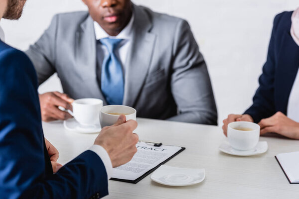 selective focus of multiethnic business partners holding cups during coffee break, cropped view