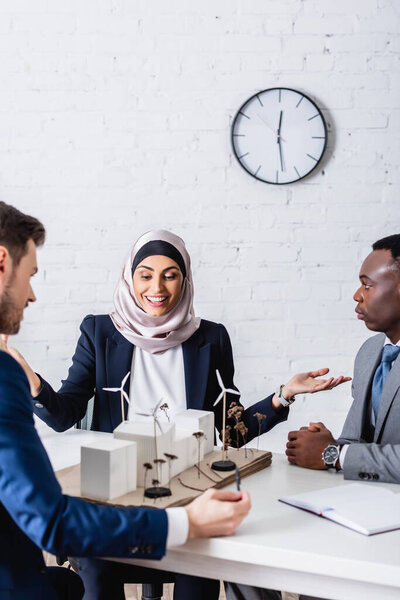 happy arabian businesswoman with open arms near green power station and multiethnic business partners, blurred foreground
