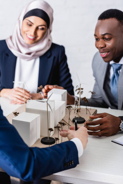 happy multicultural business partners looking at maquette of green power station, blurred background