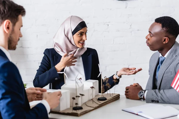 緑色の発電所のモデルの近くでアフリカ系アメリカ人のビジネスパートナーと話をしながら手を指してアラビア人のビジネス女性を笑顔で — ストック写真