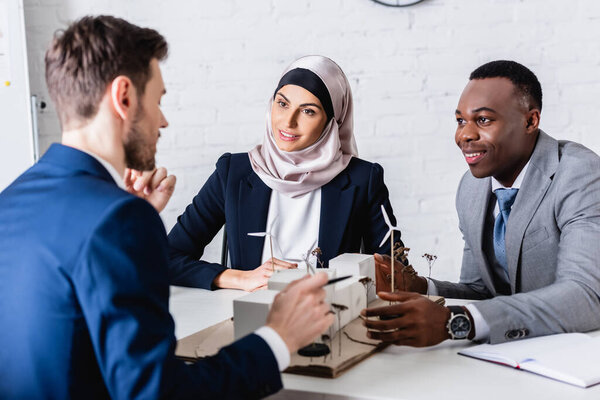 smiling arabian and african american business partners looking at businessman near model of alternative power station, blurred foreground  