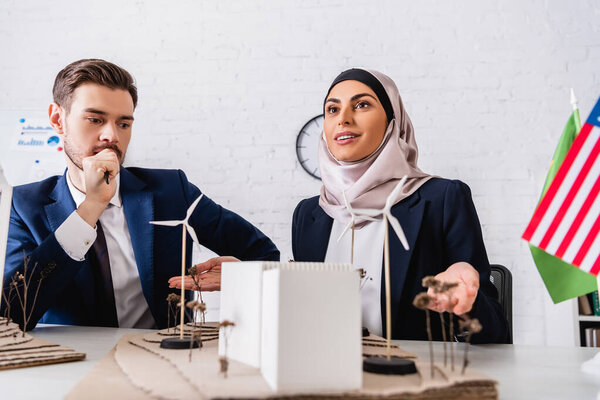 smiling arabian businesswoman pointing with hands at model of alternative power station near business partner, blurred foreground