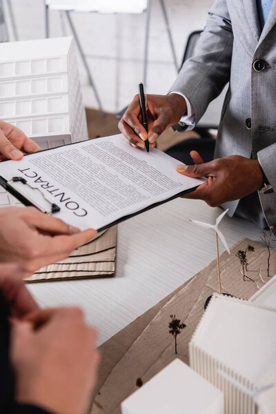 partial view of african american businessman signing contract near green power station model and business partners of blurred foreground