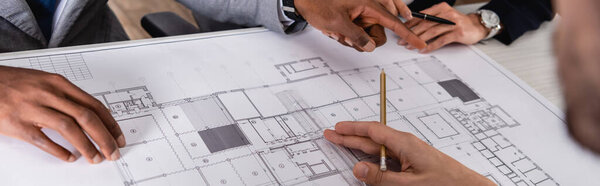 cropped view of african american man pointing with finger near blueprint and business partners, blurred foreground, banner