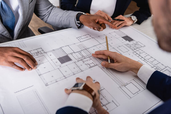 partial view of african american businessman pointing with finger at blueprint near multicultural partners, blurred foreground
