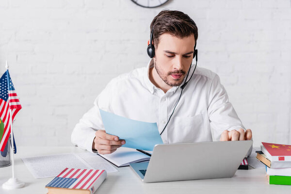 young translator in headset holding document while working on laptop near dictionaries and american flag 