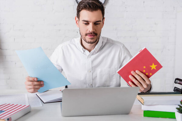 smiling interpreter holding chinese dictionary and document near laptop in translation agency