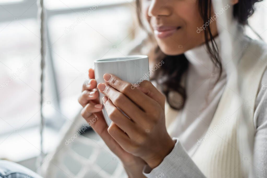 partial view of young african american woman holding cup of warm tea at home, blurred background