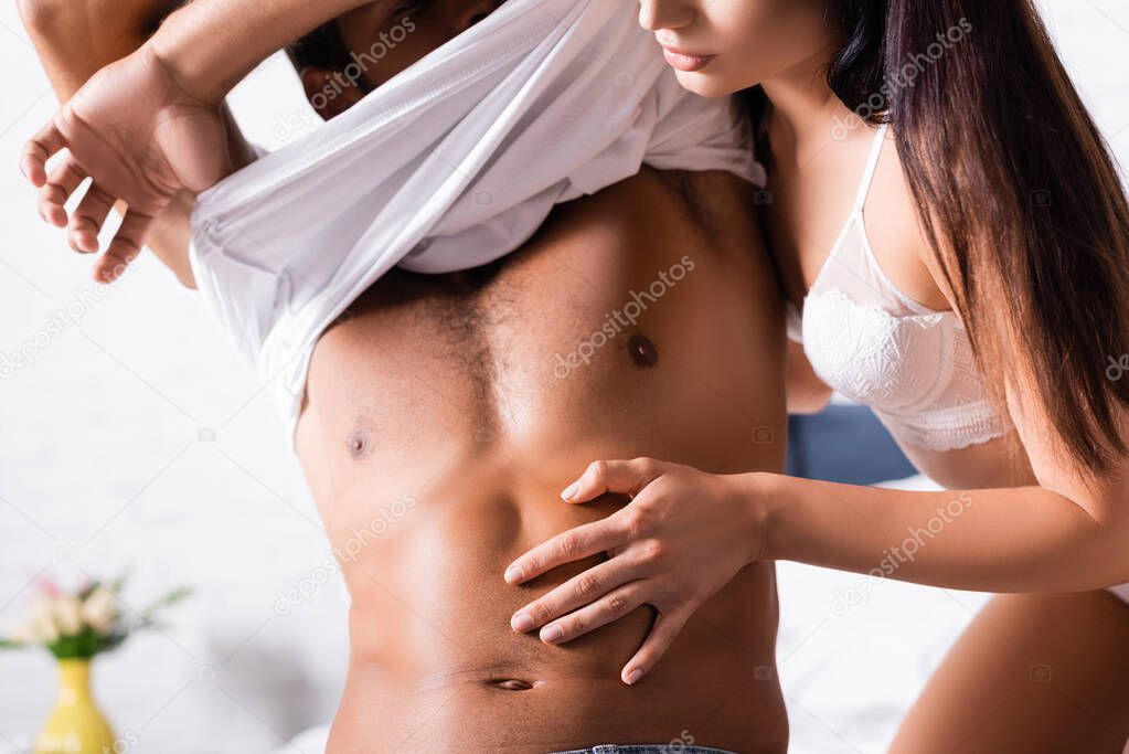 cropped view of seductive woman in underwear touching torso of man undressing on blurred background indoors