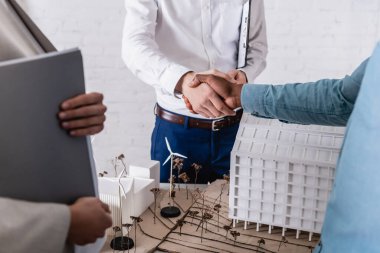 partial view of african american businessman shaking hands with business partner near alternative power station maquette, blurred foreground clipart