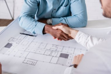 cropped view of african american businessman shaking hands with business partner near blueprint, blurred foreground clipart