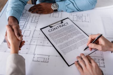 cropped view of businessman signing contract near interracial partners shaking hands near blueprint  clipart