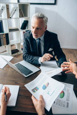 Ofiste dizüstü bilgisayarın yanında elinde belgelerle iş adamlarına bakan olgun bir yatırımcı. 