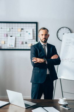 Ciddi işadamı kameraya bakıyor. Kolları çapraz. Cihazların yanında ve ön planda bulanık defter var. 