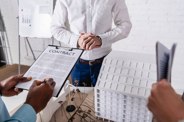 cropped view of african american businessman signing contract near model of green power station and business partners, blurred foreground