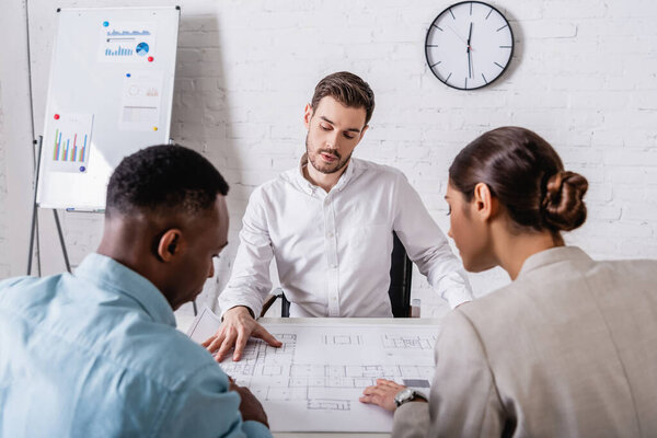 multicultural businesspeople looking at blueprint near interpreter and flipchart with infographics on background