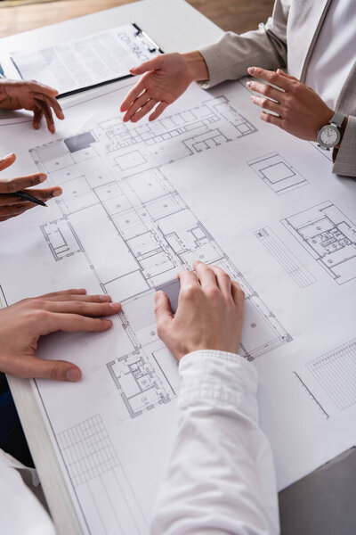 cropped view of businesswoman pointing with hand at blueprint near multiethnic business partners