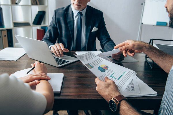 Cropped view of businessman pointing at papers with charts near colleague with notebook and investor using laptop on blurred background 