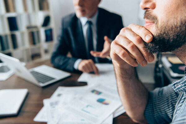 Cropped view of businessman with hand near chin sitting near investor and papers on blurred background in office 