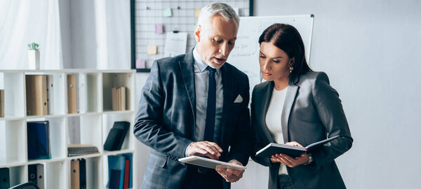 Mature investor using digital tablet near businesswoman with notebook in office, banner 