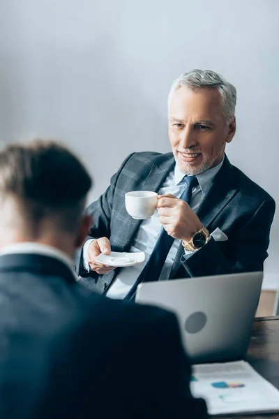 Positiver Investor Hält Tasse Kaffee Neben Laptop Und Geschäftsmann Verschwommenen — Stockfoto